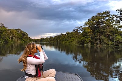 A traveler in a life vest scans the riverbank with binoculars from the bow of a skiff during a wildlife tour on a calm Amazon river lined with rainforest, Brazil.