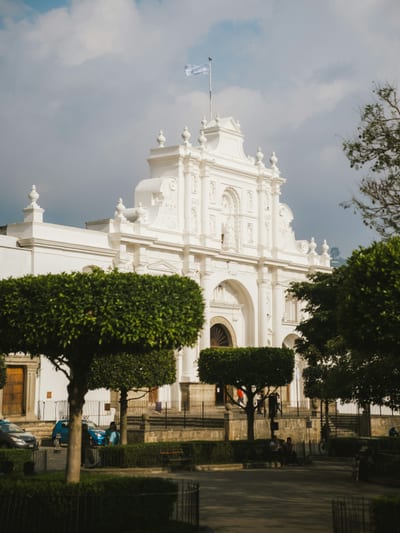 The white baroque facade of the Antigua Cathedral rising over the manicured topiary of the Parque Central in Antigua, Guatemala.
