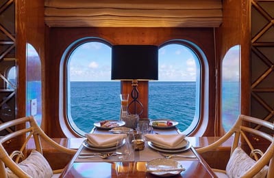 A wood-paneled dining nook aboard the Aqua Mare luxury yacht is set for a meal beside a large window framing the open ocean of the Galápagos Islands, Ecuador.