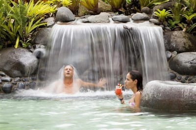 A couple relaxing under a rock waterfall in a hot-spring pool, the woman sipping a tropical cocktail, at Arenal Springs Resort in La Fortuna, Costa Rica