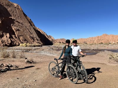A couple in helmets stand with two mountain bikes on a dry riverbed trail framed by towering red-rock canyon walls in the Atacama Desert near San Pedro de Atacama, Chile.