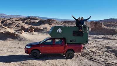 A cheering couple sits on the roof of a red pickup with a green overland camper unit, arms raised, parked amid the eroded rock formations of the Atacama Desert near San Pedro de Atacama, Chile.