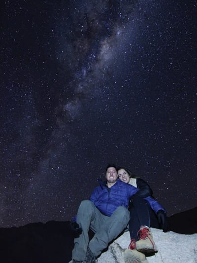 A couple sits together on a rock beneath the bright arc of the Milky Way in the clear, star-filled night sky of the Atacama Desert, Chile.