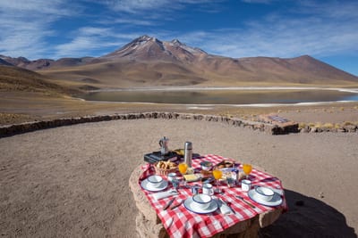 An outdoor breakfast picnic with a red-checkered cloth, coffee, juice and pastries set on a stone table overlooking a still altiplano lagoon and snow-dusted volcanic peak in the high Atacama Desert near San Pedro de Atacama, Chile.