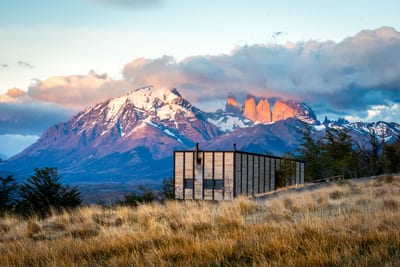A wood-clad private villa at Awasi Patagonia lodge stands in golden grassland beneath the snow-capped peaks and granite towers of Torres del Paine at sunset, Chile.