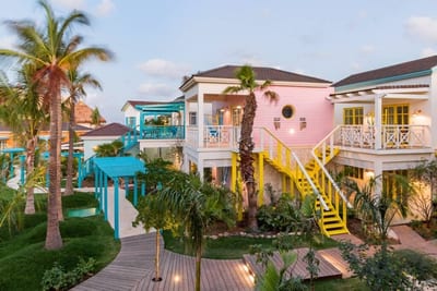 Pastel pink, turquoise, and yellow two-level cottages with palm trees, lush lawns, and a winding boardwalk at dusk at Boardwalk Boutique Hotel, Aruba.