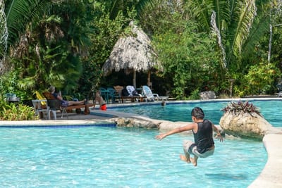 A boy leaps mid-air into a bright blue free-form pool at a boutique jungle lodge, with a thatched palapa, lounge chairs, and lush tropical palms surrounding the water in Belize.