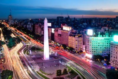 Aerial twilight view over Buenos Aires showing the Obelisco rising from the wide Avenida 9 de Julio, with car light trails streaking through the city as the sky glows pink at sunset, Argentina.