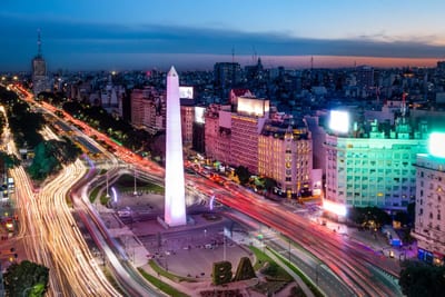A twilight aerial of Buenos Aires showing the white Obelisco rising from the wide Avenida 9 de Julio, with red and white car light trails streaking through the avenues, a lit "BA" sign, and a pink sunset over the skyline, Argentina.