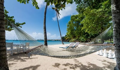 A string hammock between palm trees on a white-sand beach at Calabash Luxury Boutique, with sun loungers, a giant chess set, and a turquoise bay dotted with sailboats behind, Grenada.