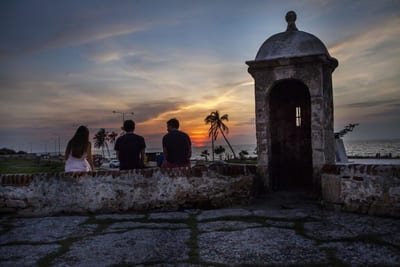 Three people sit on the colonial stone city walls of Cartagena beside a domed sentry box, watching the sun set over the Caribbean Sea behind silhouetted palms, Colombia.