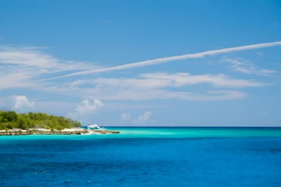 A moored white boat sits off the green, white-sand shoreline of Catalina Island amid bands of turquoise and deep-blue Caribbean water under a bright sky, Dominican Republic.