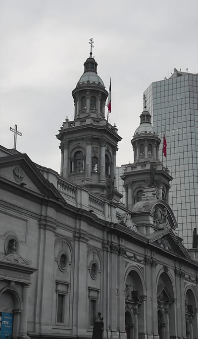 A historic cathedral with ornate twin towers topped by domes and crosses, each flying the Chilean flag. The stone façade features arched windows and intricate carvings, with a modern glass skyscraper rising in the background under a cloudy sky.