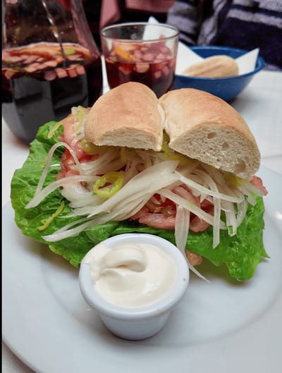 A large Chilean fish sandwich on a soft roll, topped with lettuce, tomato, and shredded green beans, served with a ramekin of mayonnaise at Liguria restaurant in Santiago, Chile.
