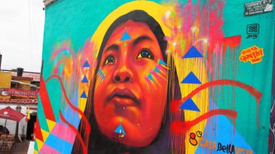 A close-up of a colorful Comuna 13 mural depicting an indigenous girl's face with blue triangles and dripping paint on a turquoise wall in Medellín, Colombia.