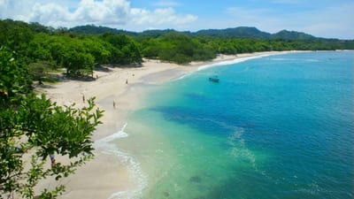 A long crescent of pale-gold sand fringed by dense green forest meets calm turquoise water with a small boat anchored offshore on a Guanacaste beach, Pacific coast of Costa Rica.