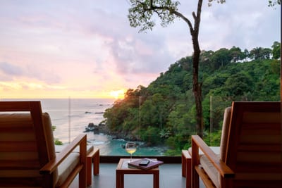 A premium suite terrace with wooden lounge chairs and a glass of wine framing a sunset over a forested cove and the Pacific Ocean in Costa Rica.