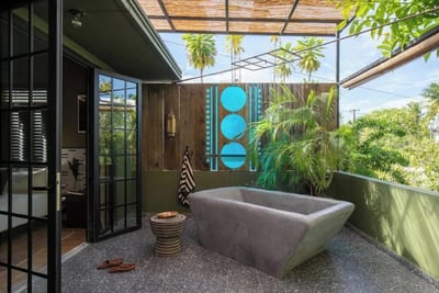 A sculptural stone soaking bathtub on a private tropical terrace with a carved wooden privacy screen and palms at Dreamcatcher by DW, Curaçao.
