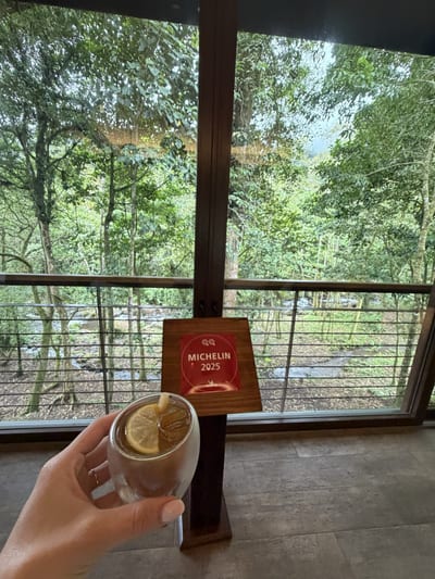 A welcome drink held up in front of the Michelin 2025 Key plaque at El Silencio Lodge, with the river and cloud forest behind, Bajos del Toro, Costa Rica