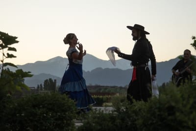 A folk dancer in a layered blue skirt and a gaucho in a wide-brimmed hat performing at dusk with a guitarist and the Andes behind, at Entre Cielos, Vistalba, Mendoza, Argentina