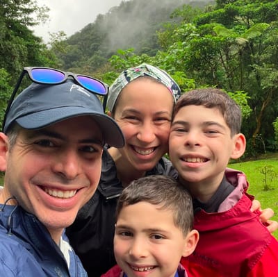 A smiling family of five takes a selfie on a misty cloud-forest trail in Costa Rica, with green mountains and low-hanging clouds behind them during a guided hike.
