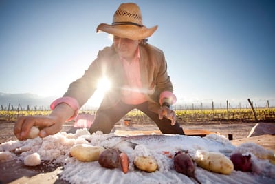 Chef Francis Mallmann in a straw hat packing coarse salt over potatoes, carrots and beets on an outdoor table beside the vineyard rows at Siete Fuegos, The Vines Resort, Mendoza, Argentina