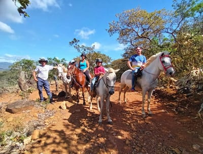 A group of visitors on horseback ride along a mountain trail with green hills behind them at Guayabo Animal Rescue &amp; Sanctuary, Costa Rica.