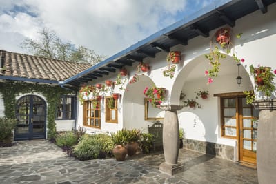 A whitewashed colonial hacienda courtyard with arched stone columns, a terracotta-tiled roof and hanging red geraniums opens onto a cobblestone patio at Hacienda Zuleta in the Ecuadorian Andes.