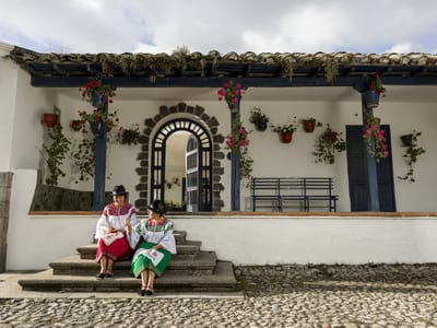 Two Otavaleña women in traditional embroidered blouses, hats and colorful wrap skirts sit doing handwork on the stone steps of the whitewashed colonial veranda at Hacienda Zuleta in the Ecuadorian Andes.