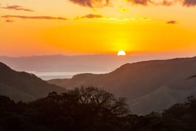 The sun sets behind layered mountain ridges in Costa Rica's highlands, with silhouetted cloud-forest treetops in the foreground and a distant gulf shimmering in the golden light.