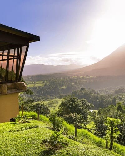 A glass-walled hilltop villa overlooks rolling green hills toward the cone of the Arenal Volcano in Costa Rica, with warm sunlight breaking over the ridge in the Arenal/La Fortuna region.