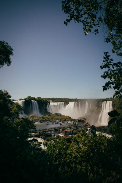A daytime view of the Iguazú Falls cascades with a boardwalk crossing the river, framed by lush jungle, Brazil.
