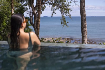 A woman relaxes in a villa infinity pool overlooking the Pacific Ocean and distant islands, framed by tropical trees at Isla Palenque private island, Panama.