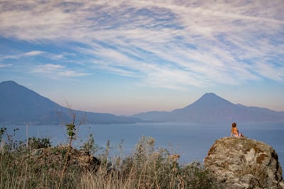 A traveler sits on a rock above the calm water of Lake Atitlán, the volcanoes rising across the lake at golden hour, Guatemala.