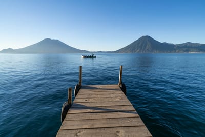 Lake Atitlán at sunrise with traditional wooden boats moored along a pier, ringed by three volcanic peaks rising from the highland water.