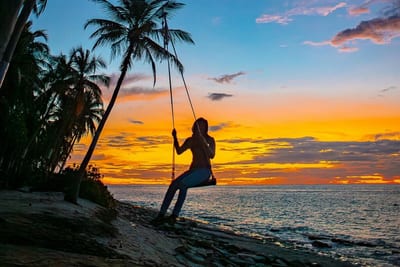 A traveler sits silhouetted on a wooden rope swing strung from a leaning palm tree on the beach as the Caribbean sky glows orange at sunset on Little Corn Island, Nicaragua.