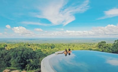 A couple relaxes at the edge of a luxury infinity pool overlooking an endless rainforest canopy stretching to the horizon under a bright blue sky at a hilltop lodge in Costa Rica.
