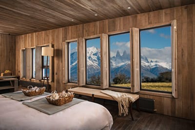 Wood-panelled luxury lodge suite framing the snow-capped granite peaks of Torres del Paine through panoramic windows, Chilean Patagonia.