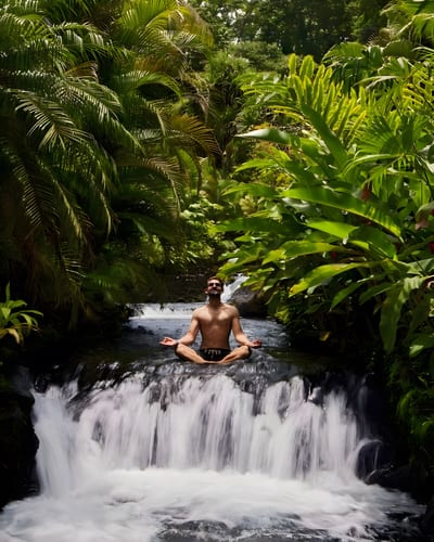 Man sitting cross-legged in meditation atop a small waterfall surrounded by lush rainforest in Costa Rica