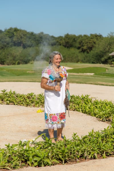 A woman in a hand-embroidered Yucatecan dress holds a smoking copal incense burner during a traditional Mexican blessing ceremony on the manicured grounds of a golf resort, Mexico.