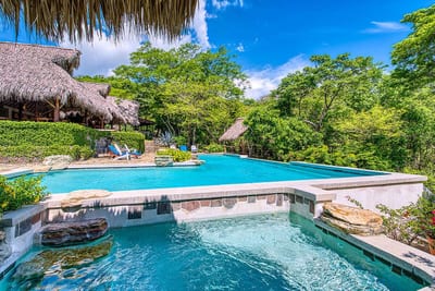 A multi-level swimming pool with a stone hot tub, thatched palapas and sun loungers set among lush tropical jungle under a blue sky at Morgan's Rock eco-lodge, Nicaragua.