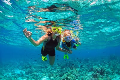A mother and child snorkel together just below the surface of clear turquoise water, both in masks and bright fins giving an OK sign, with a coral reef and small fish below on the Belize Barrier Reef.