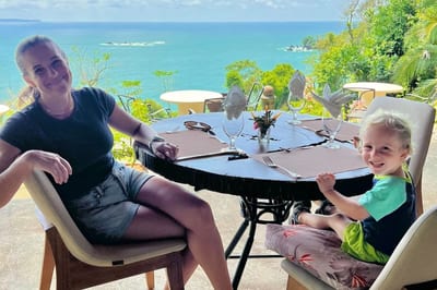 A smiling mother and her young son sit at a set table on an open-air restaurant terrace overlooking the ocean and a forested coastline in Manuel Antonio, Costa Rica.