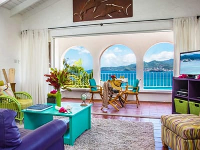 A colorful villa living room with arched openings framing a turquoise bay and green hills at Mount Cinnamon Beach & Wellness Resort, Grenada.