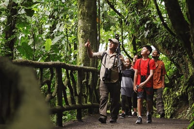 A naturalist guide with a spotting scope points up into the rainforest canopy as a family of visitors watches during a guided wildlife tour in Costa Rica, looking for sloths, monkeys, and birds.