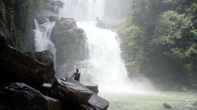 A powerful multi-tier cascade plunges into a misty plunge pool as a lone visitor sits on a rock at the base of Nauyaca Waterfalls near Dominical, Costa Rica.