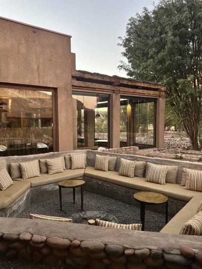 A sunken stone firepit lounge with an L-shaped banquette of striped cushions and brass side tables at the adobe Nayara Alto Atacama lodge at dusk, San Pedro de Atacama, Chile.