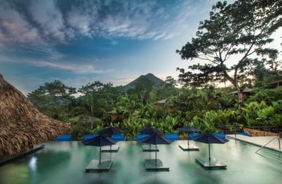 Infinity pool with submerged blue daybeds and umbrellas at Nayara Springs resort in La Fortuna, with Arenal Volcano rising above tropical jungle