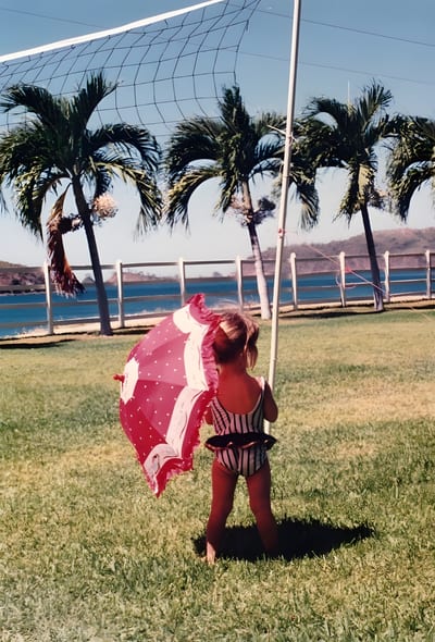 A vintage childhood photo of a young girl in a striped swimsuit holding a pink parasol on a grassy lawn, with palm trees and a blue bay behind her on Costa Rica's Nicoya Peninsula.