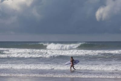 A surfer carries a white surfboard along the shoreline with waves crashing under a moody cloudy sky on the Nicoya Peninsula, Costa Rica.